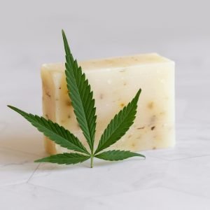 Craft soap bar near green cannabis leaves on white marble table. Cosmetic Mockup